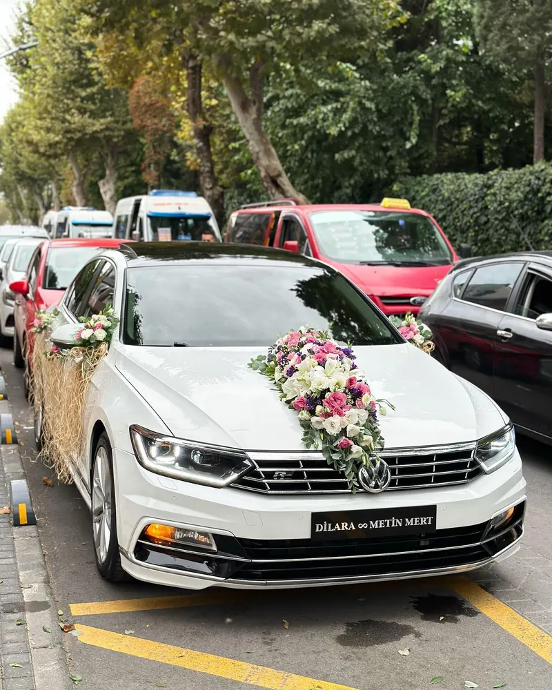 Kaputunda pembe-beyaz çiçek şeridi ve isim plakası olan beyaz sedan gelin arabası